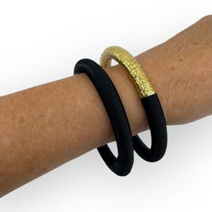 May include: Two bracelets on a wrist. One is black, the other is gold. The gold bracelet has a textured surface. The black bracelet is smooth. Both bracelets are round and have a thick band.