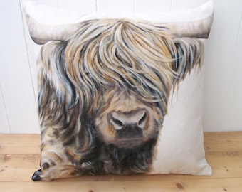 May include: A square throw pillow featuring a detailed, realistic illustration of a Highland cow with long, shaggy, golden-brown and grey fur covering its eyes. The cow has prominent curved horns and is set against a plain, off-white background.