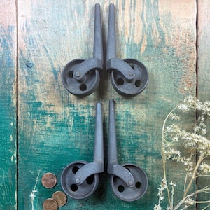 May include: Four antique metal caster wheels with a dark gray finish are arranged on a weathered, teal and brown wooden surface. The wheels have a classic design, with a round wheel and a long, tapered stem. A few pennies are scattered near the wheels.