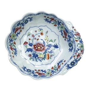 May include: A white ceramic bowl with a scalloped edge and a floral design in blue, red, and yellow. The bowl has two handles on the sides.