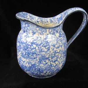 Puede incluir: Jarra de cerámica azul y blanca con un patrón moteado. La jarra tiene un asa curva y un pico ligeramente acampanado. El borde está adornado con una línea azul oscuro o negra. La jarra es probablemente vintage o antigua.