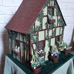 Op de afbeelding: Een miniatuurhuis met een bruin dak, groene muren en bruine randen. Het huis is versierd met nep klimop en heeft kleine bloembakken onder de ramen. Het huis staat op een groene basis.
