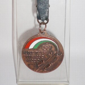 Puede incluir: Un llavero de color bronce con una correa de cuero y un dije circular. El dije presenta la bandera italiana, los anillos olímpicos y el texto "Federazione Italiana Pattinaggio Ghiaccio".
