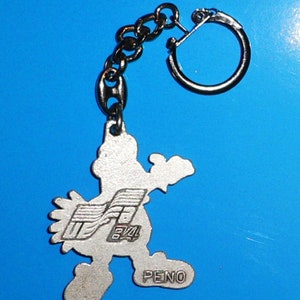 Puede incluir: Llavero de plata con un personaje de dibujos animados con un dise&ntilde;o estilizado. El personaje sostiene un logotipo con el texto "PENO" y "84" en &eacute;l.