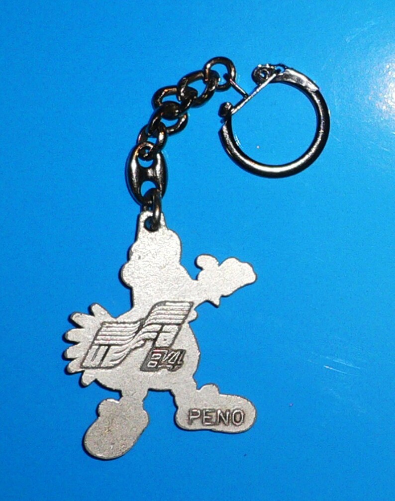 Puede incluir: Llavero de plata con un personaje de dibujos animados con un dise&ntilde;o estilizado. El personaje sostiene un logotipo con el texto "PENO" y "84" en &eacute;l.
