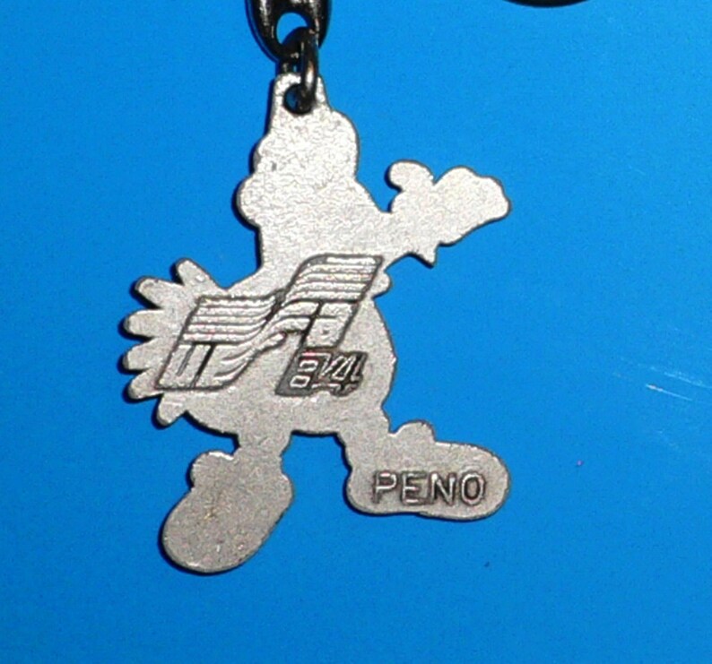 Puede incluir: Llavero de color plateado con un personaje de dibujos animados con un dise&ntilde;o estilizado y el texto "PENO" en la parte inferior.