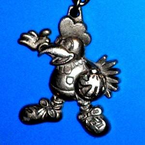 Puede incluir: Un llavero de metal plateado con forma de gallo de dibujos animados que sostiene una pelota de baloncesto. El gallo lleva una camiseta con un n&uacute;mero y tiene un pico grande.