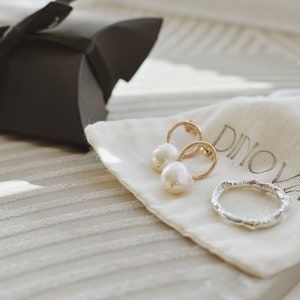 May include: A white pearl and gold hoop earring set and a silver ring on a white fabric pouch with the word "Dinova" printed on it.