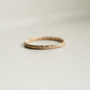 May include: A textured gold ring, shown in close-up. The ring has a detailed, irregular surface. The band is set against a neutral background. The ring is likely a wedding band or a ring for everyday wear. The ring is gold in colour.