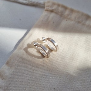 May include: A pair of silver hoop earrings with a slightly rounded, flat surface. The earrings are on a white fabric background.