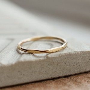 May include: A simple gold band ring with a hammered texture. The ring is sitting on a light gray textured surface.