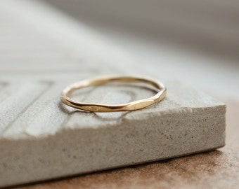 May include: A simple gold band ring with a hammered texture. The ring is sitting on a light gray textured surface.