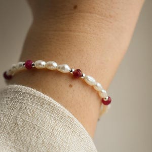 May include: A delicate bracelet featuring white, oval-shaped pearls interspersed with small, round, red beads. The bracelet is secured with a silver clasp and is worn on a wrist, against a neutral-coloured fabric.