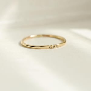 May include: A delicate gold ring with three small, evenly spaced, engraved triangle shapes. The ring is thin and has a polished finish, reflecting the light. The ring is a minimalist piece of jewellery.