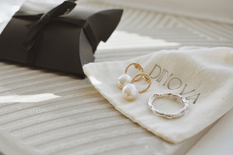 May include: A white pearl and gold hoop earring set and a silver ring on a white fabric pouch with the word "Dinova" printed on it.