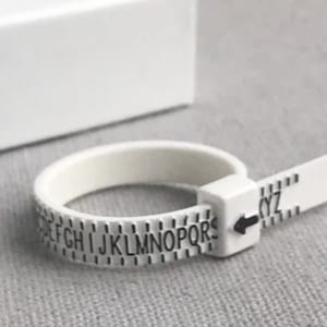 Könnte beinhalten: Ein weißer Kunststoff-Ringgrößenmesser mit einem schwarzen Pfeil und den Buchstaben A bis Z.