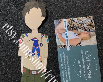 May include: A paper cutout of a boy scout figure wearing a tan shirt, green pants, and a blue neckerchief. A business card with contact information is next to the figure. The Etsy shop name and Instagram handle are visible.