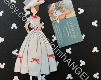 May include: A white paper doll dressed as Mary Poppins with a red bow on her dress and a red bow on her hat. The doll is standing on a black background with white polka dots.