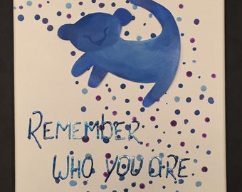 May include: A blue watercolor painting of a lion cub with the words "Remember who you are" written in silver glitter. The background is white with blue and purple polka dots.