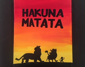 May include: A hand-painted canvas with a sunset background and black silhouettes of Simba, Pumbaa, and Timon from the Disney movie The Lion King. The text "Hakuna Matata" is written in black at the top of the canvas.
