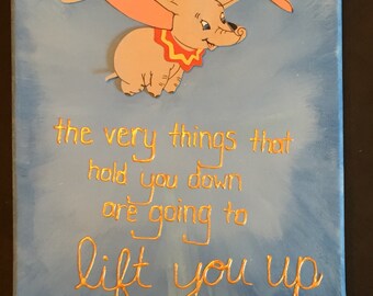 May include: A blue canvas painting with a 3D image of Dumbo the elephant flying with a small figure of Timothy Mouse on his back. The painting has gold lettering that reads "the very things that hold you down are going to lift you up".