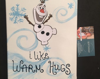 May include: A white canvas painting with a blue and white snowflake design. A paper cutout of Olaf from the movie Frozen is on the canvas. The text "i like WARM HUGS" is written in black paint on the canvas.