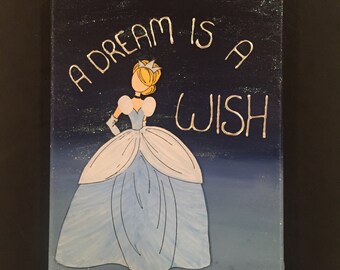 May include: A canvas painting featuring a cartoon illustration of Cinderella in a light blue gown. The text "A DREAM IS A WISH" is written in silver script above the character. The background transitions from dark blue to light blue.
