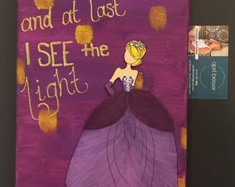 May include: A canvas painting with the words "and at last I SEE the light" in gold script. A cutout of a princess in a purple and lavender gown is on the right. Gold accents and a business card are also visible.