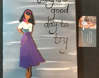 May include: A gray canvas painting with a white and gray striped background. The painting features a cutout of a woman with dark hair, a pink headband, and a purple skirt. The text "today is a good day to try" is written in black and gold lettering.