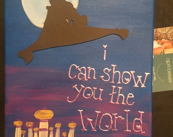 May include: A hand-painted canvas with a silhouette of Aladdin and Abu flying on a magic carpet in front of a full moon. The text "I can show you the world" is written in pink below the carpet. A white building with gold domes is painted at the bottom of the canvas.