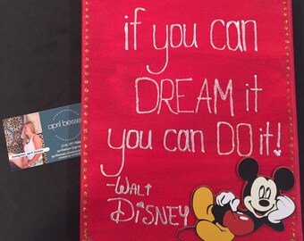 May include: A red canvas with gold glitter trim features a white handwritten quote: "if you can DREAM it you can DO it! -WALT DISNEY". A felt Mickey Mouse figure is attached to the bottom right corner.