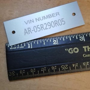 May include: A silver metal tag with the text "VIN NUMBER AR-05R290R05" engraved on it. The tag has two holes on the top and bottom.