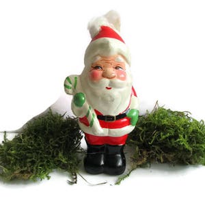 May include: A vintage Santa Claus figurine, hand-painted with a red and white hat and suit, black boots, and a white beard. He holds a green and white candy cane. Green moss is arranged around the figurine, creating a festive holiday scene.