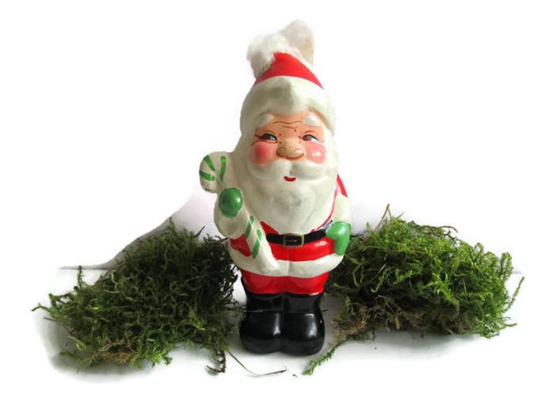 May include: A vintage Santa Claus figurine, hand-painted with a red and white hat and suit, black boots, and a white beard. He holds a green and white candy cane. Green moss is arranged around the figurine, creating a festive holiday scene.
