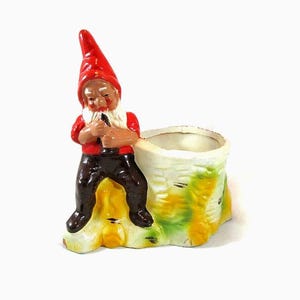 May include: A ceramic gnome figurine with a red hat, red shirt, and black pants, leaning against a tree stump planter. The planter is white with green and yellow accents. A whimsical garden decor item.