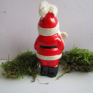 May include: A ceramic Santa Claus figurine, viewed from the back, with a red coat, black belt, and black boots. The hat is red with white trim and a fluffy white top. The figurine is on a white surface with green moss.