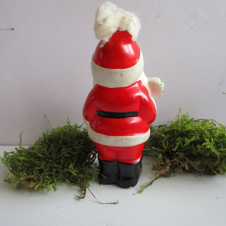 May include: A ceramic Santa Claus figurine, viewed from the back, with a red coat, black belt, and black boots. The hat is red with white trim and a fluffy white top. The figurine is on a white surface with green moss.