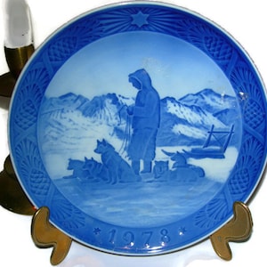 Puede incluir: Un plato de colección redondo de porcelana azul y blanca con una escena en relieve de una persona con parka con capucha junto a un equipo de trineo de perros en un paisaje montañoso nevado. El año 1978 está grabado y el borde tiene piñas.