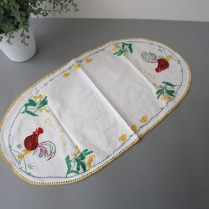 May include: White oval table runner with embroidered roosters and floral designs. The text "Glad Gask" and "Jula Pous" is embroidered in gold thread.