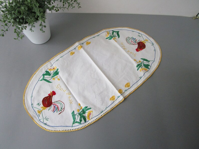 May include: White oval table runner with embroidered roosters and floral designs. The text "Glad Gask" and "Jula Pous" is embroidered in gold thread.