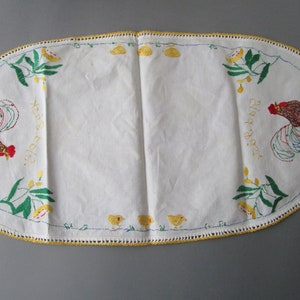 May include: White oval table runner with embroidered roosters, chicks, and leaves. The text "Happy Easter" is embroidered on each side of the runner.