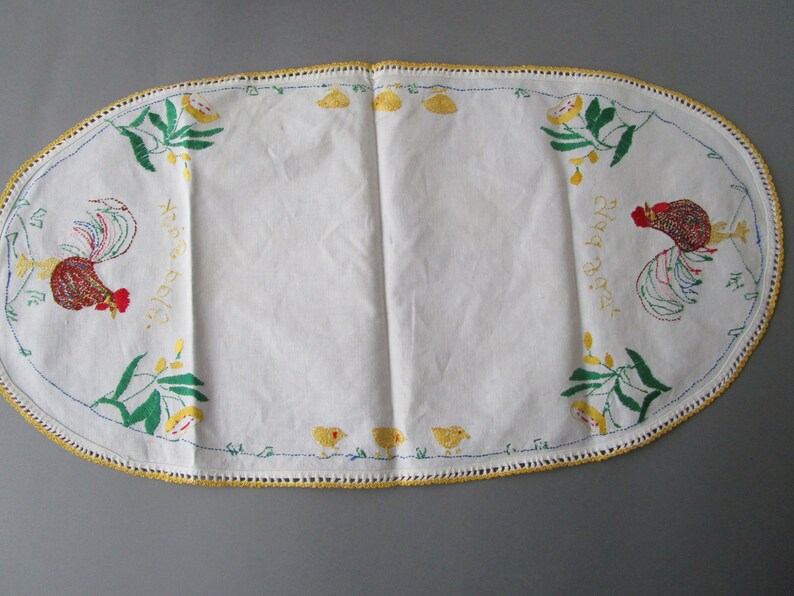 May include: White oval table runner with embroidered roosters, chicks, and leaves. The text "Happy Easter" is embroidered on each side of the runner.
