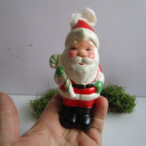 May include: A vintage Santa Claus figurine, featuring a red and white hat with a fluffy pom-pom, a white beard, and a red suit. He holds a green and white striped candy cane and wears black boots. The figurine is held in a hand.