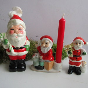 May include: A collection of vintage Christmas figurines featuring Santa Claus. The figurines are painted in red, white, and black, with details like a candy cane, a teddy bear, and a red candle. The Santa Claus figurines are in various poses.