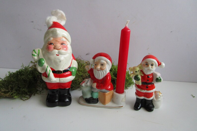 May include: A collection of vintage Christmas figurines featuring Santa Claus. The figurines are painted in red, white, and black, with details like a candy cane, a teddy bear, and a red candle. The Santa Claus figurines are in various poses.