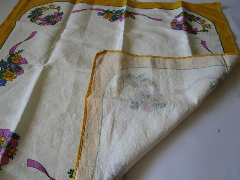 May include: A white tablecloth with a yellow border and floral designs. The tablecloth is folded in half, revealing the underside with a similar floral design.