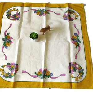 May include: A white tablecloth with a yellow border and floral designs. The tablecloth has a repeating pattern of wreaths with pink ribbons and flowers.