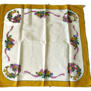 May include: A white tablecloth with a yellow border and floral designs. The floral designs feature pink ribbons, yellow flowers, and green leaves.