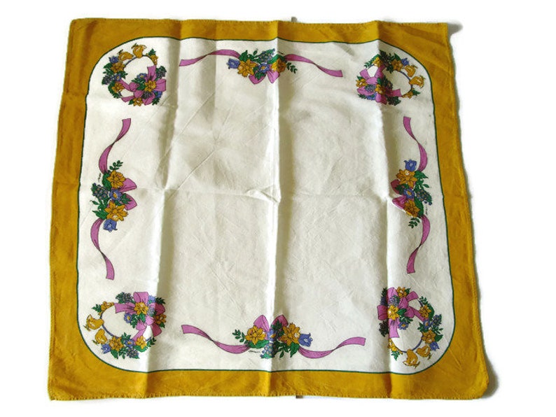 May include: A white tablecloth with a yellow border and floral designs. The floral designs feature pink ribbons, yellow flowers, and green leaves.