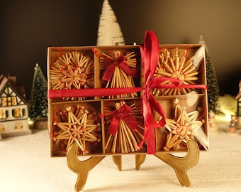 Pode incluir: Um conjunto de seis enfeites de Natal de palha numa caixa, amarrados com uma fita vermelha. Os enfeites incluem estrelas e anjos. A caixa é exibida em um suporte dourado, com casas e árvores em miniatura ao fundo.